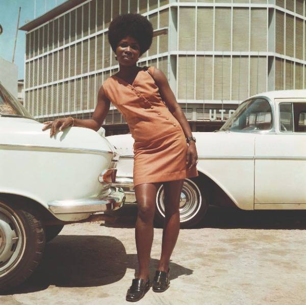 A woman wearing an orange mini dress leans on a white car as she poses outside the United Trading Company headquarters in Accra, Ghana, 1971