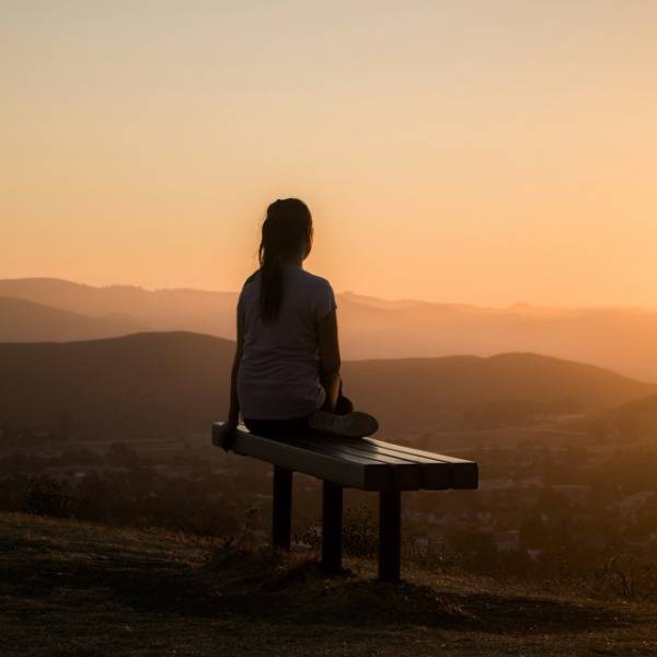 A woman sits on a bench at the top of a hill, looking out over the view at sunset