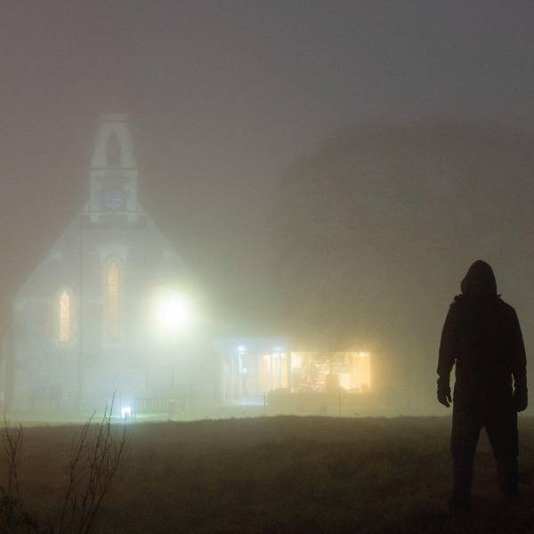 A man whose face cannot be seen stands outside a church in the dark