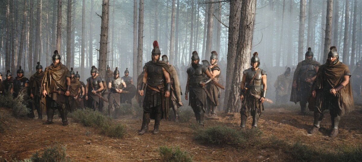 Odysseus and his men, dressed in armour, stand together in a forest
