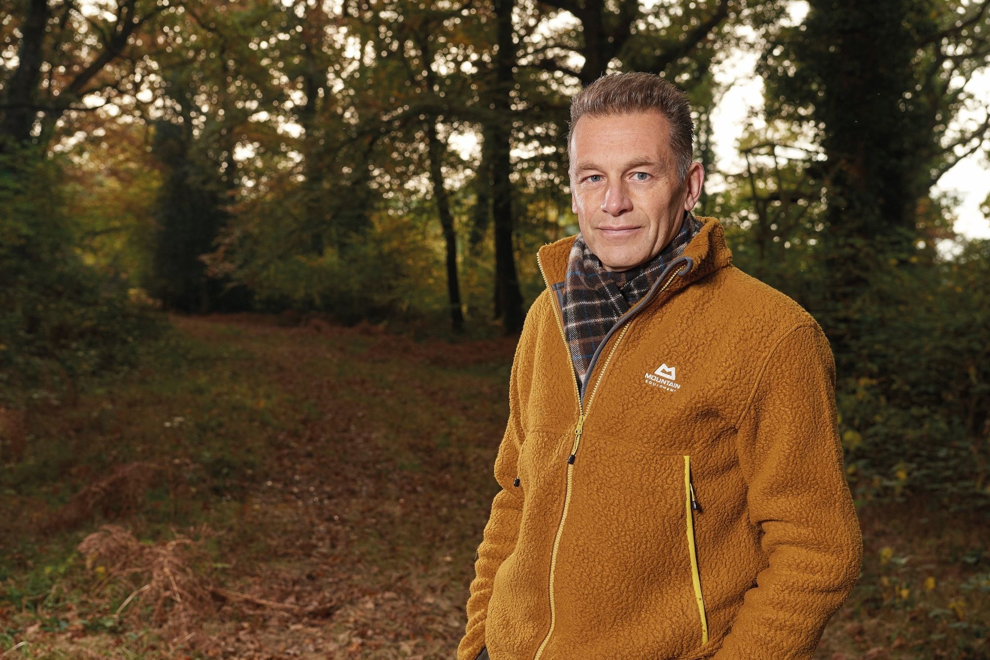 Chris Packham stands in the woods wearing a fleece jacket and scarf