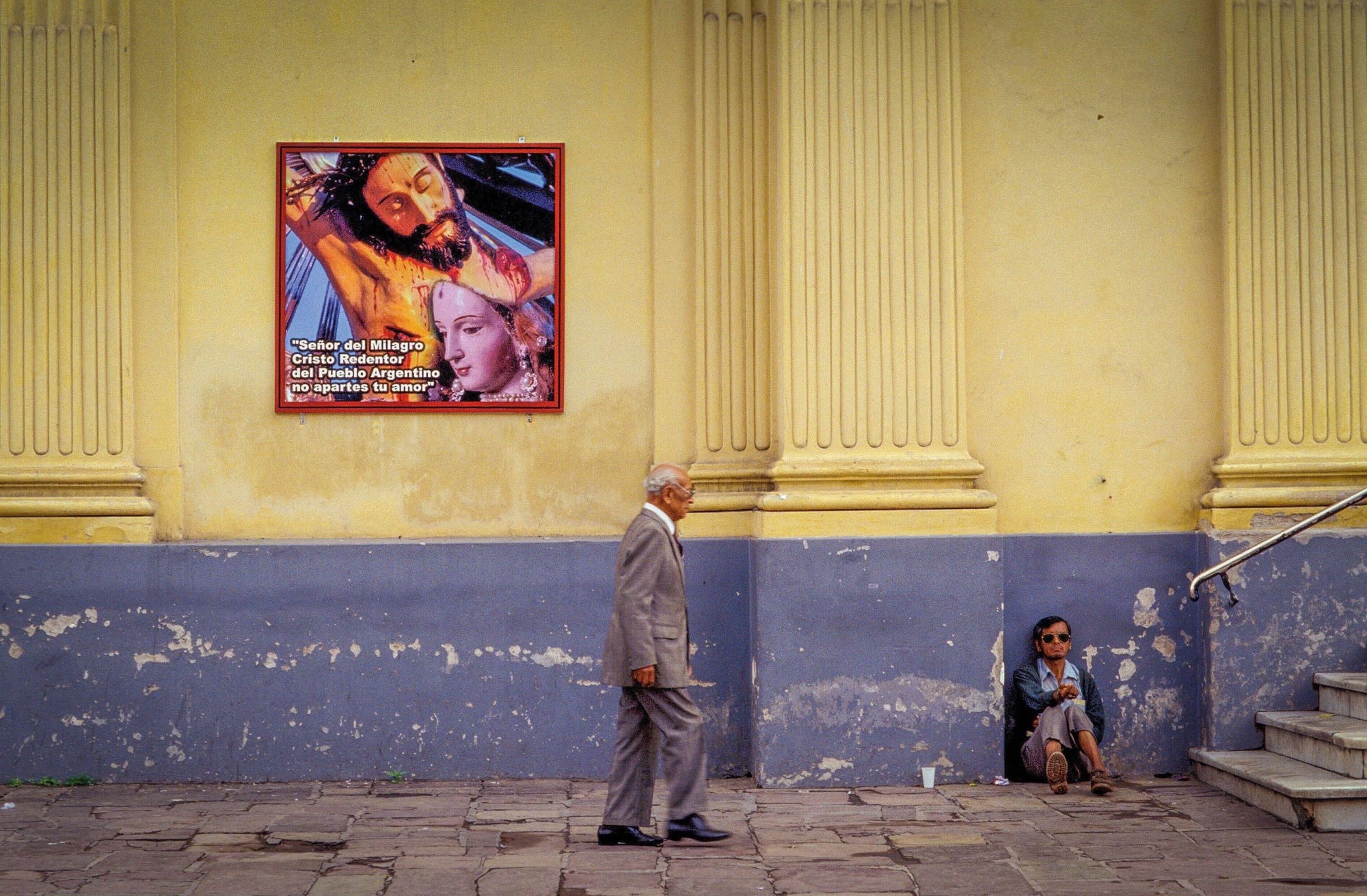 A man sits on the pavement begging for money as a man in a suit walks by, next to the cathedral in Salta, Argentina