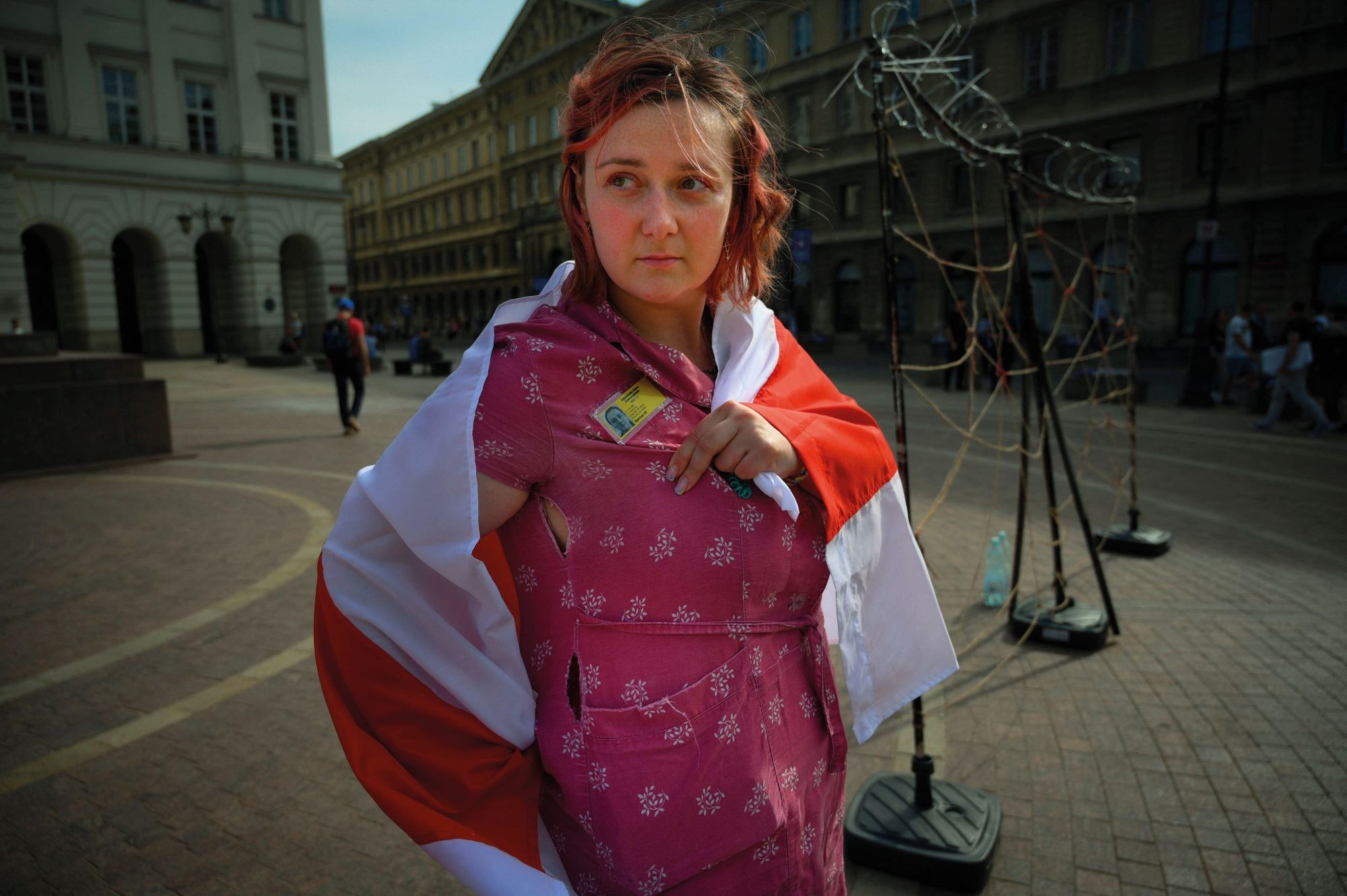 Darya Afanasyeva in Warsaw, Poland, where she has received asylum, displaying her political prisoner yellow tag. She is draped in the historic red-and-white flag of Belarus, which is still used by opposition groups today