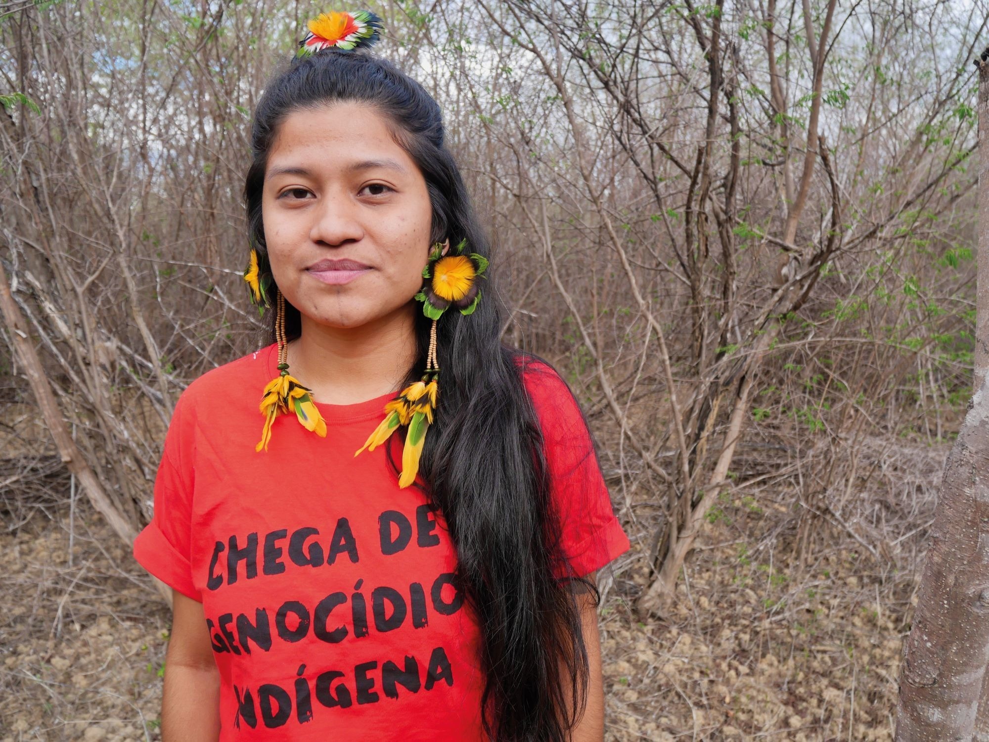 Ytxaha Pankararu Pataxó wearing long feather earrings and a red T-shirt bearing the words “End the Indigenous genocide”