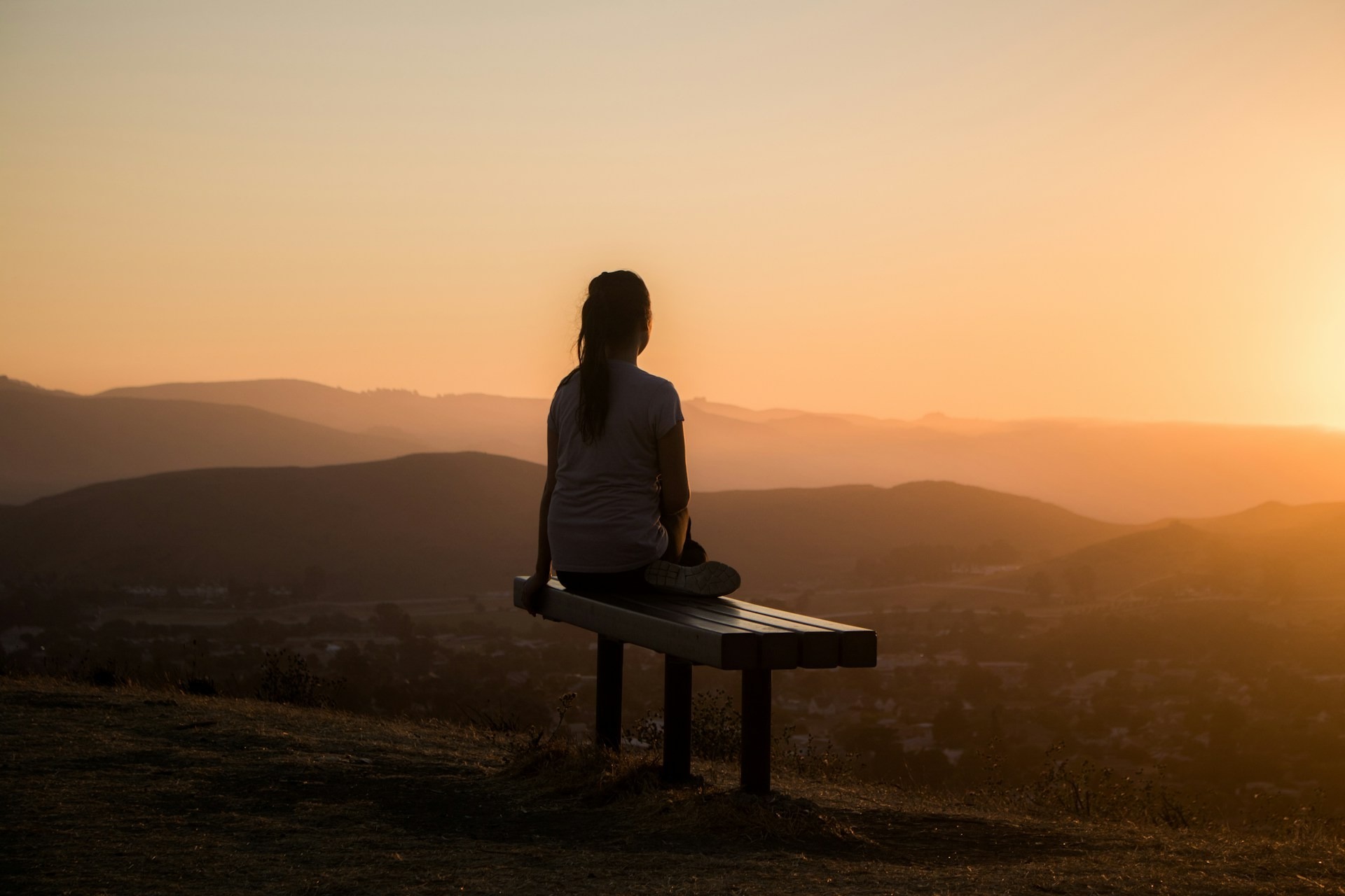 A woman sits on a bench at the top of a hill, looking out over the view at sunset